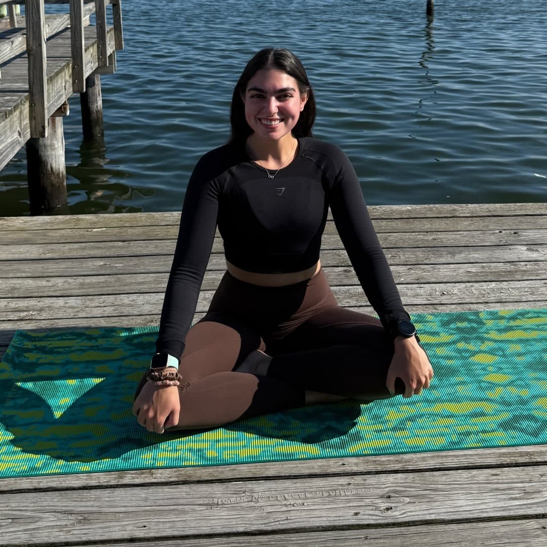 Person seated on a yoga mat on a wooden dock beside calm water on a sunny day